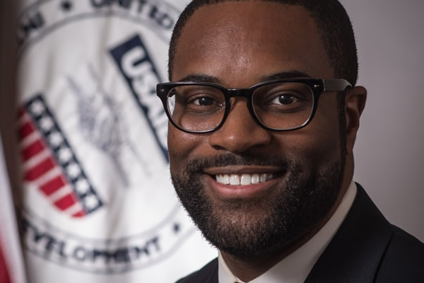 Portrait of Jay Gilliam. He is wearing a suit, tie, and glasses, and he is smiling. In the background stands a flag that bears the emblem of the United States Agency for International Development.