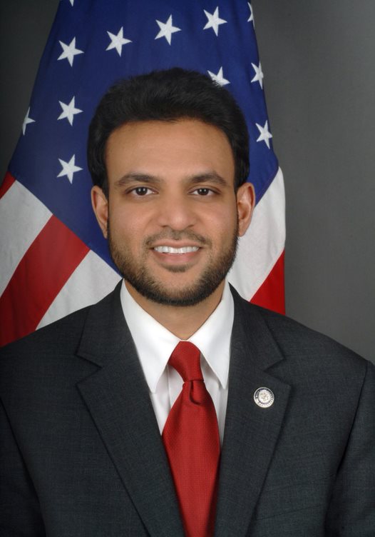 Front-facing headshot of Rashad, who is smiling and wearing a suit and tie in front of a U.S. flag.
