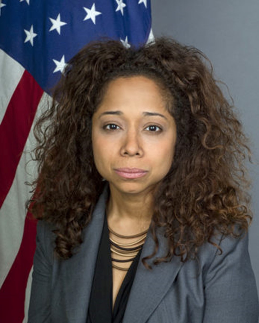 A headshot photo of Julissa Reynoso. She is wearing a grey suit with a black shirt underneath. Her hair is down and she is looking directly into the camera. There is an American flag behind here, and a grey background.
